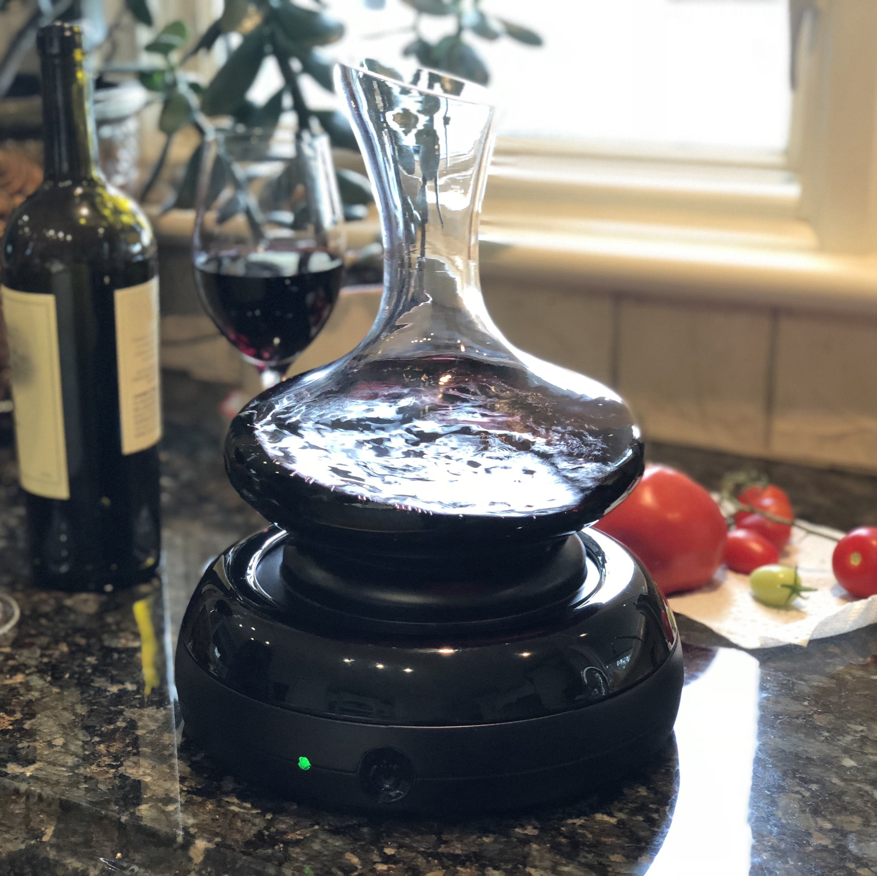 Aerisi Wine Aerator and glass decanter filled with red wine sitting on dark granite kitchen counter by white window sill. Bottle of wine, wine glass woth wine and chelrry tomatoes sitting o the counter in the background near the wine decanter.